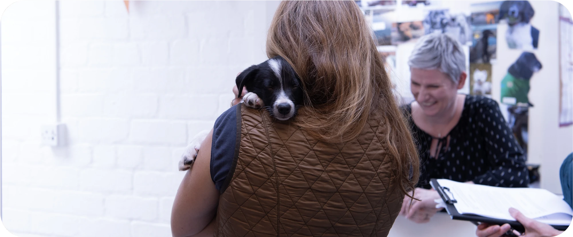 A woman with long hair and a brown vest cradles a black and white puppy on her shoulder.